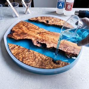 May include: A circular silicone mold containing three pieces of natural olive wood with intricate grain patterns, being filled with a translucent blue liquid epoxy resin poured from a clear plastic cup, with resin bottles and a heat gun nearby.