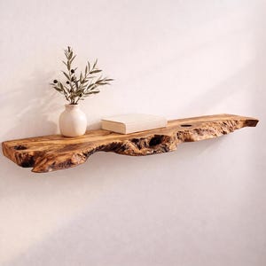 May include: A live edge wooden wall shelf with a natural brown grain and irregular bark-covered front edge. A small, cream-colored ceramic vase containing a green olive branch and a closed, rectangular beige book sit on the shelf surface.