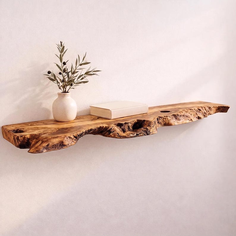 May include: A live edge wooden wall shelf with a natural brown grain and irregular bark-covered front edge. A small, cream-colored ceramic vase containing a green olive branch and a closed, rectangular beige book sit on the shelf surface.