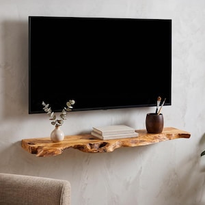 Peut inclure: Une étagère flottante en bois à bord brut fixée au mur sous un téléviseur à écran plat. L'étagère supporte un petit vase blanc avec des tiges d'eucalyptus, deux livres empilés de couleur claire et un gobelet en bois sombre avec deux pinceaux.