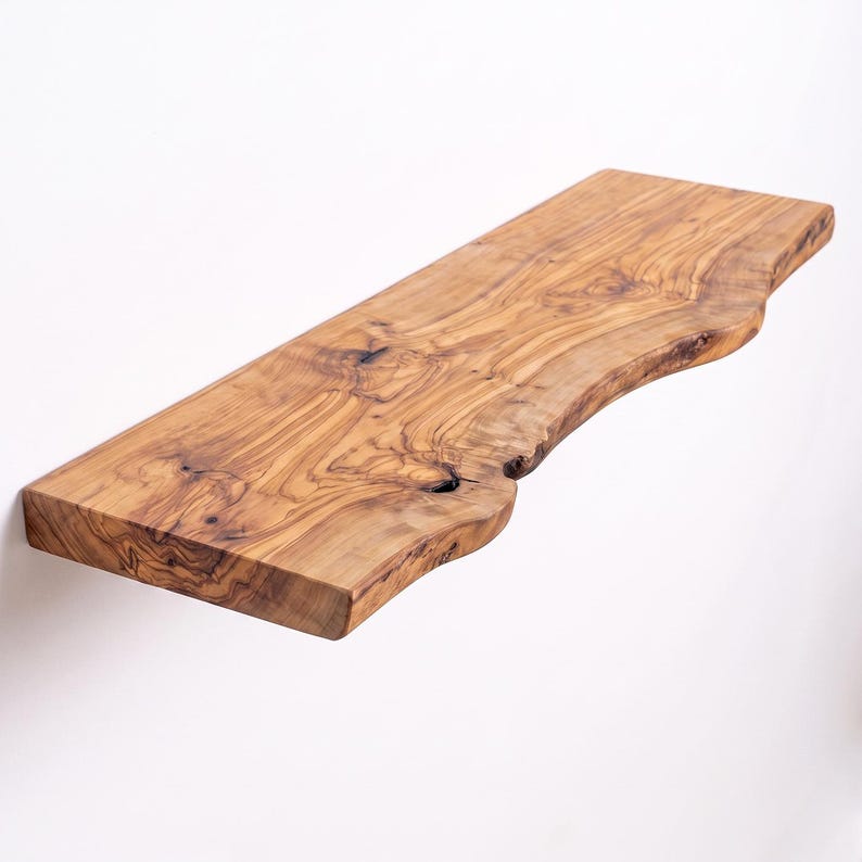 May include: A rectangular wall shelf crafted from natural olive wood, featuring a prominent live edge on the front side. The wood displays a rich, swirling grain pattern with dark brown knots and light golden-brown tones throughout the surface.