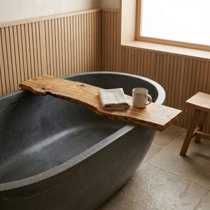 Puede incluir: Una bañera ovalada gris oscuro con una bandeja de baño de madera. La bandeja contiene una toalla blanca doblada y una taza blanca. El baño tiene paneles de madera clara y un pequeño taburete de madera.