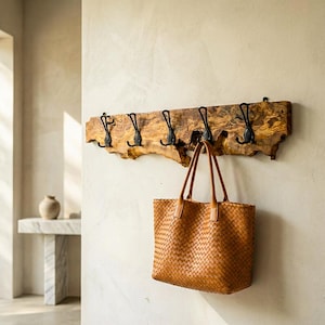 May include: A rustic wooden coat rack with six black metal hooks. A woven brown leather tote bag hangs from one of the hooks. The rack is mounted on a neutral-colored wall, creating a warm, inviting entryway.