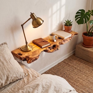 May include: A live edge wooden floating shelf mounted on a wall, featuring a natural wood grain and irregular bark edges. The shelf holds a brass desk lamp, a brown leather-bound journal, a ceramic mug, and a closed book with a light cover.