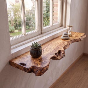 Puede incluir: Una repisa de madera de borde natural con veteado visible y un borde frontal irregular cubierto de corteza. Sobre la repisa hay una pequeña suculenta en una maceta de cerámica oscura y tres piedras grises lisas junto a un reloj de arena.