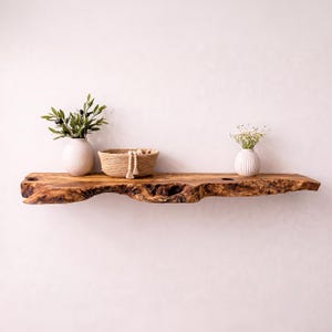 May include: A live edge wooden floating shelf with a natural grain pattern. On the shelf sits a white textured vase with green olive branches, a woven jute basket containing wooden beads, and a small white ribbed vase with white flowers.