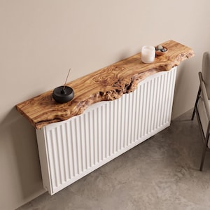Puede incluir: Una estantería de madera rectangular de borde natural con veta visible y borde frontal irregular, montada sobre un radiador blanco de costillas verticales. Un porta-incienso negro y una vela blanca descansan sobre la madera.