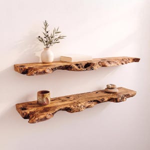 May include: Two wall-mounted shelves made from natural live-edge wood with a prominent grain. The top shelf holds a small white vase with an olive branch and a book. The bottom shelf features a small ceramic cup and two stacked stones.