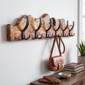 May include: A wall-mounted coat rack featuring a natural live-edge wood plank with a prominent grain pattern. It includes six matte black metal hooks, each with a curved design and a textured base. A brown leather bag hangs from one hook.