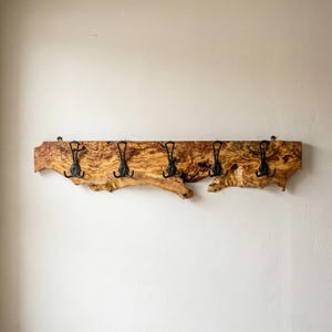 Può includere: Un attaccapanni rustico in legno con bordo naturale. Il legno presenta un motivo a venature marrone e beige caldo. Cinque ganci in metallo nero sono attaccati, offrendo una soluzione di archiviazione funzionale e decorativa per cappotti e cappelli.