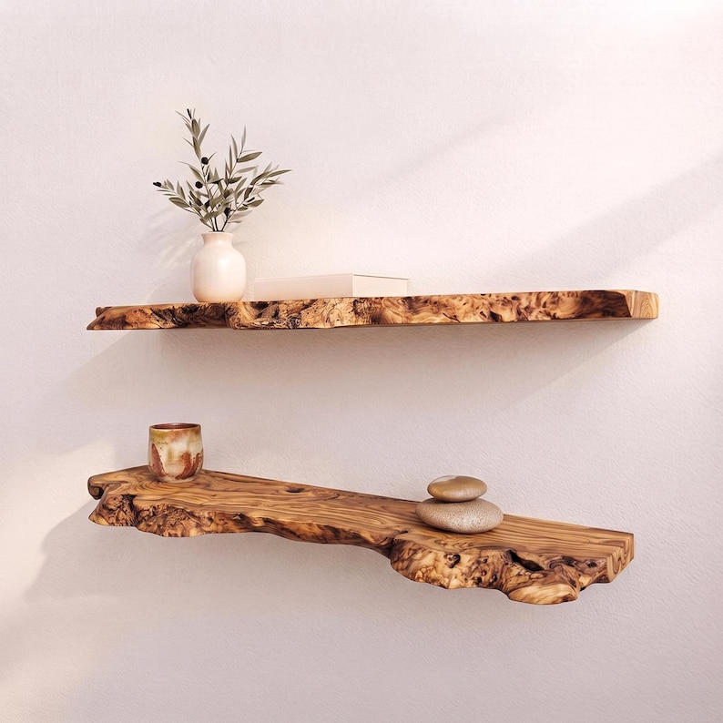 May include: Two horizontal wall shelves made from natural wood with irregular live edges. The top shelf holds a white vase with olive branches and a book. The bottom shelf holds a small patterned ceramic cup and two stacked smooth stones.