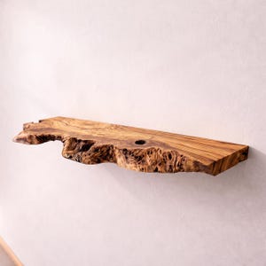 May include: A wall-mounted shelf crafted from a single piece of natural olive wood. The shelf features a prominent live edge with a rough, textured bark-like appearance and a smooth, polished top surface showing distinct wood grain patterns.