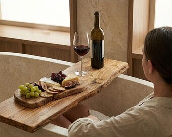 May include: A live-edge wooden bathtub tray resting across a stone bathtub. The tray holds a glass of red wine, a dark glass wine bottle, and a wooden serving board topped with green grapes, red grapes, sliced figs, cheese, and crackers.