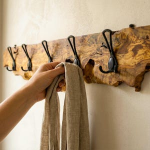 May include: A wooden coat rack with five black metal hooks. The wood has a natural grain with brown and tan tones. A hand is holding a beige scarf, ready to be hung. The rack is mounted on a light-coloured wall. The rack's length is approximately 60 cm.