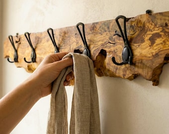 May include: A wall-mounted coat rack featuring a natural live-edge wood plank with prominent grain patterns and five matte black metal hooks. The wooden board has an irregular, organic shape and is mounted horizontally on a plain wall.