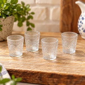May include: A set of four clear glass tumblers featuring a textured exterior with a repeating circular medallion pattern, vertical fluting along the base, and a beaded rim detail. These small drinking vessels are made of transparent glass.