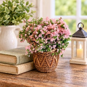 May include: A small, round, brown woven basket planter filled with dense, bushy pink flowers and green foliage. The planter sits on a wooden surface next to two stacked, weathered, light-colored books and a small, white, square-shaped lantern.