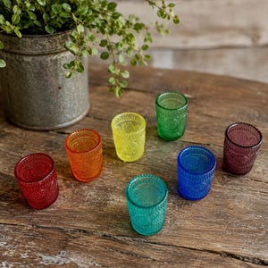 May include: A set of seven small glass votive holders in a variety of colors including red, orange, yellow, green, teal, blue, and purple. Each glass features a textured, embossed pattern on the exterior surface and a cylindrical shape.