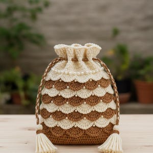 Może przedstawiać: Szydełkowa torba ze sznurkiem w kolorze kremowym i brązowym, ozdobiona perłami i frędzlami. Torba ma wzór w kształcie muszli i plecione zamknięcie ze sznurkiem. Torba stoi na drewnianej powierzchni.