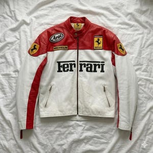 May include: A white and red leather jacket with the Ferrari logo and text. The jacket has a stand-up collar, zipper closure, and multiple patches, including "Arai" and "Michelin". The sleeves are white with red stripes.