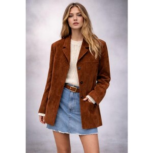 May include: A brown suede blazer with a classic lapel collar and two-button closure. The blazer is paired with a white sweater, a denim skirt, and a brown belt. The outfit is a stylish combination of textures and colors.
