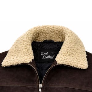 Puede incluir: Primer plano de una chaqueta de ante marrón con cuello de borrego color crema. La chaqueta tiene cierre de cremallera y una etiqueta negra que dice "Real Leather". El interior de la chaqueta es negro y acolchado.