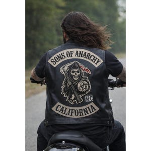 以下が含まれることがあります： 「SONS OF ANARCHY」と「CALIFORNIA」の白い文字が入った黒い革ベスト。大鎌を持った死神と頭蓋骨の大きなパッチが付いています。その人はバイクに乗っています。