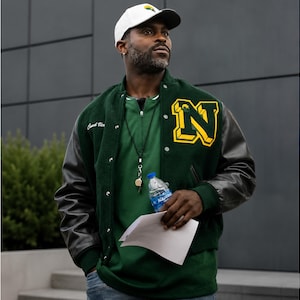 May include: A dark green varsity jacket with black leather sleeves, a large yellow and black letter N on the chest, and white snap buttons. The jacket features the text Coach Vic embroidered on the right side and is worn over a green shirt.
