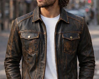 May include: A distressed brown leather jacket with two chest pockets and a collared neckline. The jacket has a zipper closure and a worn, vintage appearance. The jacket is worn over a white t-shirt.