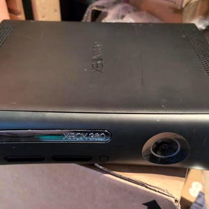 May include: Black Xbox 360 console. The console has a rectangular shape with a curved front. The front features a disc tray, power button, and the Xbox 360 logo. The top of the console has the Xbox logo. The console is sitting on a cardboard box.