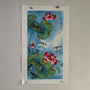 May include: A watercolor painting of lotus flowers and birds against a blue sky. The artwork features red lotus blossoms, large green lily pads, and small birds. The painting is mounted on white paper and held in place with colorful push pins.