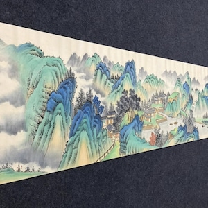 以下が含まれることがあります： 青と緑の山々、霧のかかった雲、松の木が描かれた長い横型の掛け軸。手前の小道には、赤や青の服を着た小さな人物が歩いており、川や伝統的な建物が描かれています。