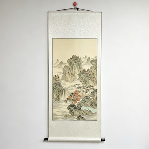 May include: A traditional Asian scroll painting depicting a mountainous landscape with a waterfall. The artwork features muted tones of green, beige, and brown, with a cream-colored border and top and bottom rollers. The scroll is hanging on a white wall.