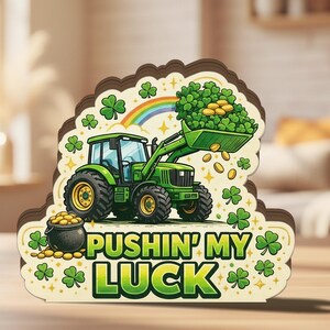 May include: A wooden St. Patrick's Day decoration featuring a green tractor scooping up gold coins and shamrocks. The text "PUSHIN' MY LUCK" is displayed below the tractor, with a rainbow and additional shamrocks. The decoration is on a wooden surface.