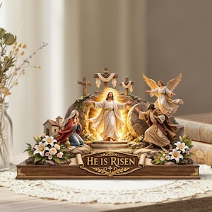 May include: A religious figurine depicting the resurrection of Jesus. The scene includes Jesus with outstretched arms, an angel, and other figures. The base is wooden, and the text "HE IS RISEN" is displayed.