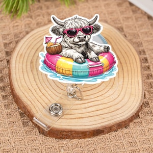 Puede incluir: Un portatarjetas con un dibujo de una vaca de las Tierras Altas con gafas de sol rosas flotando en un flotador inflable colorido. La vaca sostiene una bebida de coco con un paraguas pequeño. El portatarjetas tiene una correa de plástico transparente y un clip de metal.