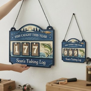 Puede incluir: Dos letreros de registro de pesca de madera azul colgados en una pared. El letrero más grande dice "FISH CAUGHT THIS YEAR" y "Sean's Fishing Log" con el número 177. El letrero más pequeño muestra el número 099.