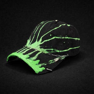 Puede incluir: Gorra de béisbol negra con diseño de salpicaduras de pintura verde neón. La gorra presenta una cadena plateada y un imperdible. Un accesorio único y artístico. La gorra es un artículo de moda elegante.