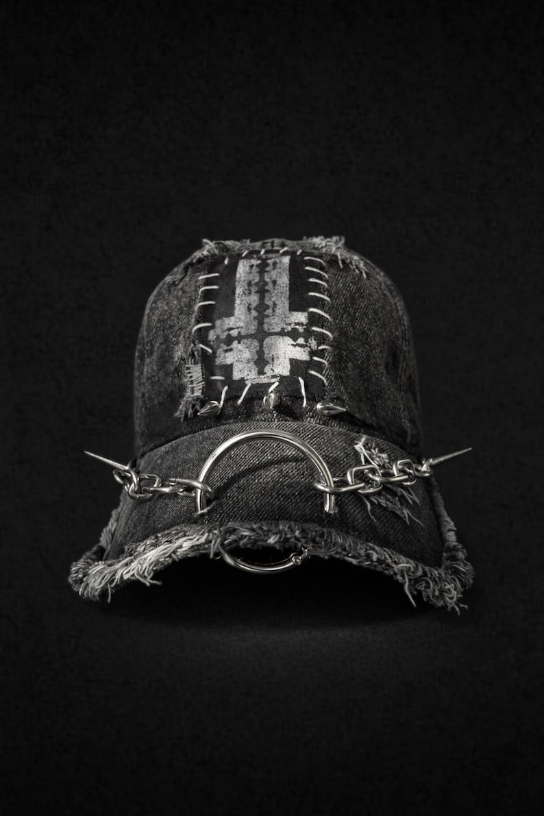 May include: A black denim baseball cap with a silver inverted cross patch and chain detailing. The cap has a distressed, frayed brim and is adorned with a silver ring and chain, and metal spikes.