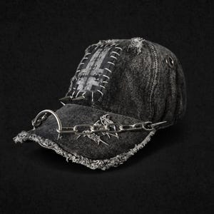 May include: Distressed black denim baseball cap with a silver chain and spike accents. The cap features a patch with white stitching and a metal ring on the side. The brim has a frayed edge, adding to the edgy aesthetic.