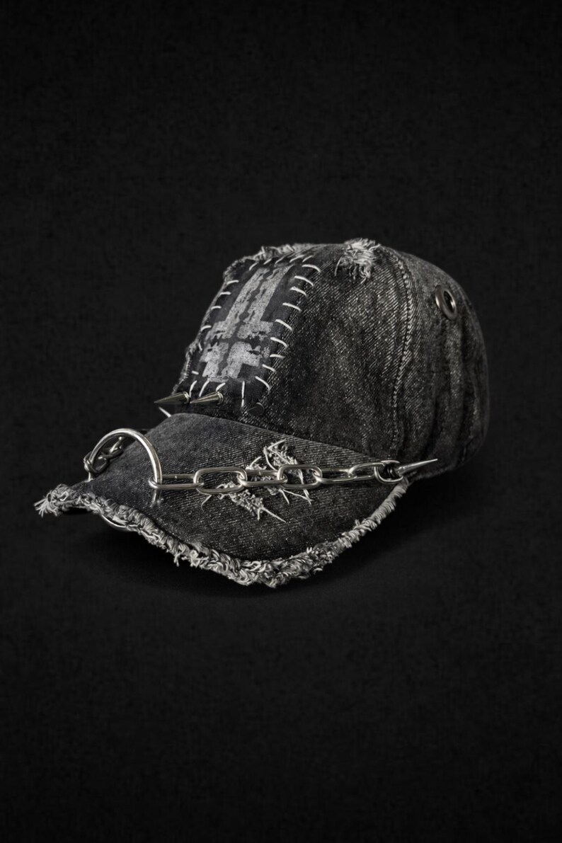May include: Distressed black denim baseball cap with a silver chain and spike accents. The cap features a patch with white stitching and a metal ring on the side. The brim has a frayed edge, adding to the edgy aesthetic.