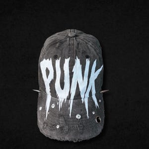Könnte beinhalten: Eine verwaschene anthrazitfarbene Baseballkappe mit dem Wort PUNK in großen, weißen, tropfenden Buchstaben auf der Vorderseite. Die Kappe ist mit silbernen Metallnieten, kleinen runden Ösen und zwei spitzen Dornen an den Seiten verziert.