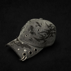 Könnte beinhalten: Graue Baseballkappe mit Used-Look, Metallspitzen und Ösen. Die Kappe hat ein Stammesdesign auf der Seite und einen gebogenen Schirm. Die Mütze ist dunkelgrau.