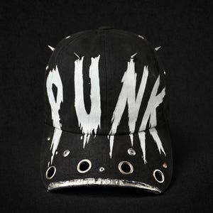 Può includere: Cappellino da baseball nero con la scritta "PUNK" in bianco, con lettere invecchiate. Il cappellino presenta occhielli in metallo argentato, una tesa argentata e punte argentate. Lo sfondo è nero.