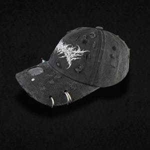 Può includere: Un cappellino da baseball nero invecchiato con una grafica ricamata bianca sul pannello frontale. Il cappellino presenta diversi anelli in metallo argentato fissati sulla visiera, insieme a un piccolo dettaglio in rete metallica e strappi.