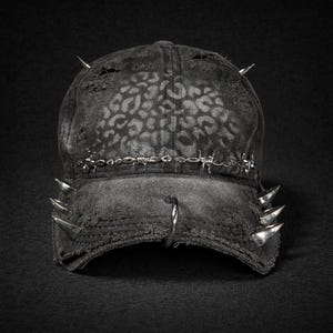 May include: A distressed black baseball cap with a leopard print pattern. The cap features silver spikes and barbed wire detailing, along with a silver hoop on the brim. The hat has a worn, edgy aesthetic.