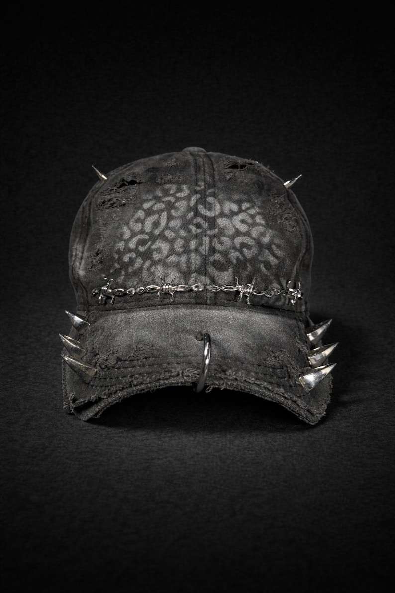 May include: A distressed black baseball cap with a leopard print pattern. The cap features silver spikes and barbed wire detailing, along with a silver hoop on the brim. The hat has a worn, edgy aesthetic.