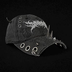Könnte beinhalten: Eine schwarze Baseballkappe im Used-Look mit silbernen Metallspitzen am Schirm und an der Seite, silbernen Metallösen und einer dünnen silbernen Kette auf der Vorderseite. Die Kappe zeigt weißen, gestickten Text in einer gezackten, stilisierten Schrift.
