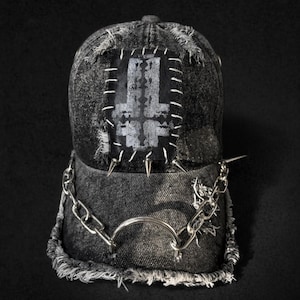 May include: A distressed denim baseball cap with a patch featuring a gray inverted cross design. The cap is embellished with silver-colored spikes, a chain, and a metal ring. The brim and edges have a frayed appearance.