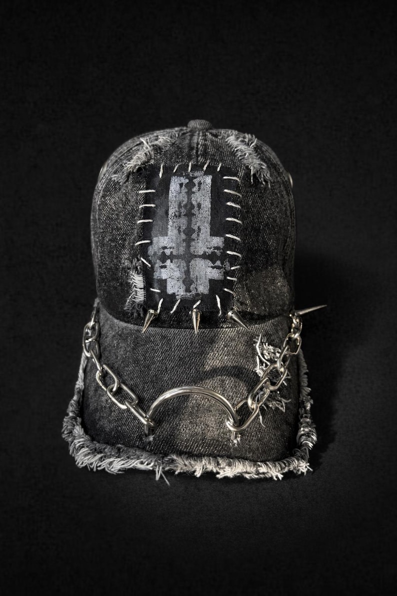 May include: A distressed denim baseball cap with a patch featuring a gray inverted cross design. The cap is embellished with silver-colored spikes, a chain, and a metal ring. The brim and edges have a frayed appearance.